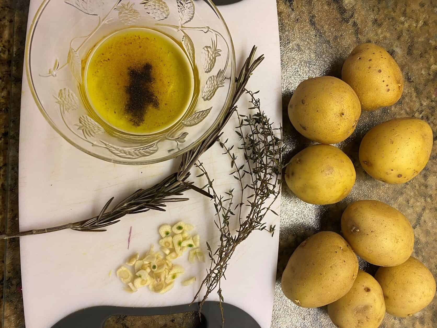 olive oil, salt peper in a dish, herbs on a cutting board and potatoes