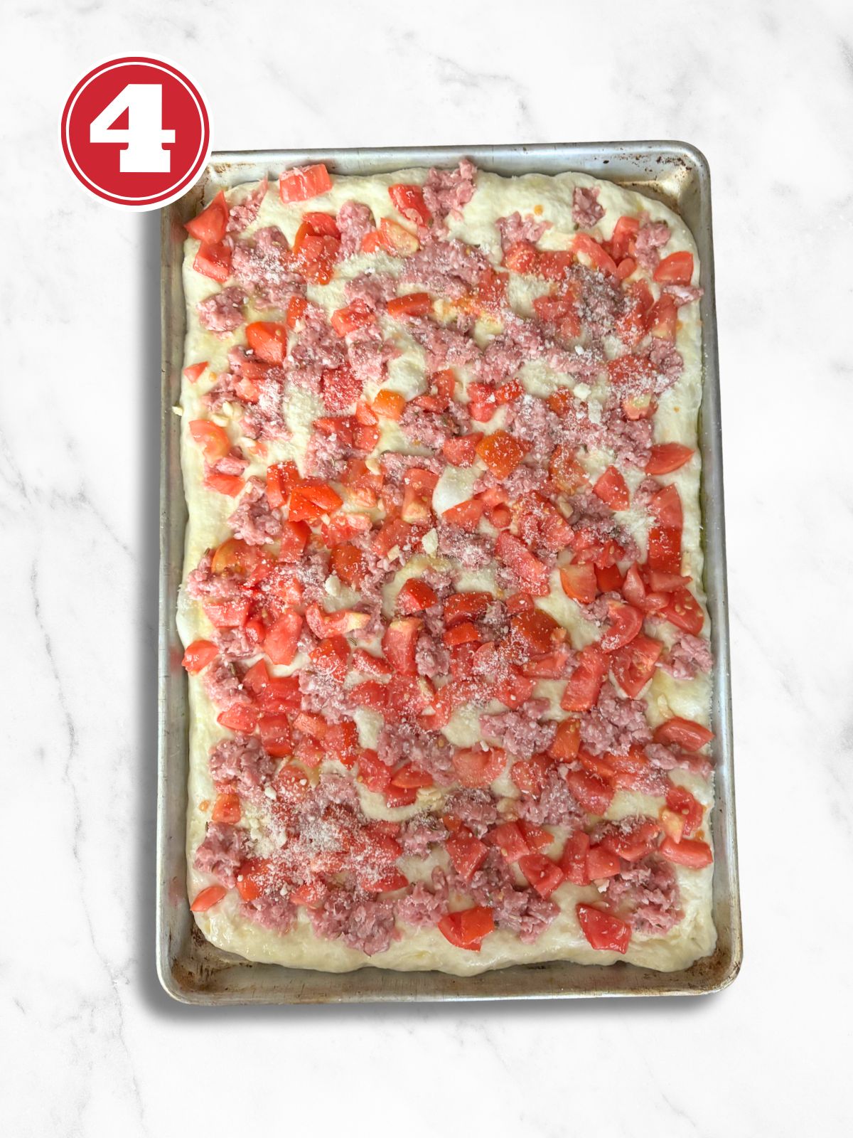 pizza dough in an aluminum pizza tray topped with sausage and diced tomato